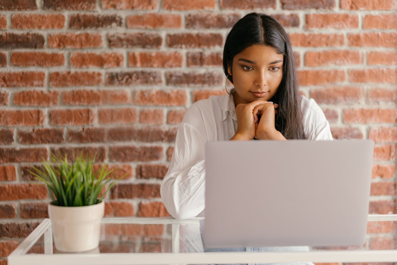 Girl with black hair in a white shirt in front of a laptop at a desk.